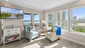 Habitación con un ventanal grande que ofrece vistas a la playa y edificios altos. Hay un televisor montado en la pared, un pequeño tocador blanco y dos sillones con cojines.