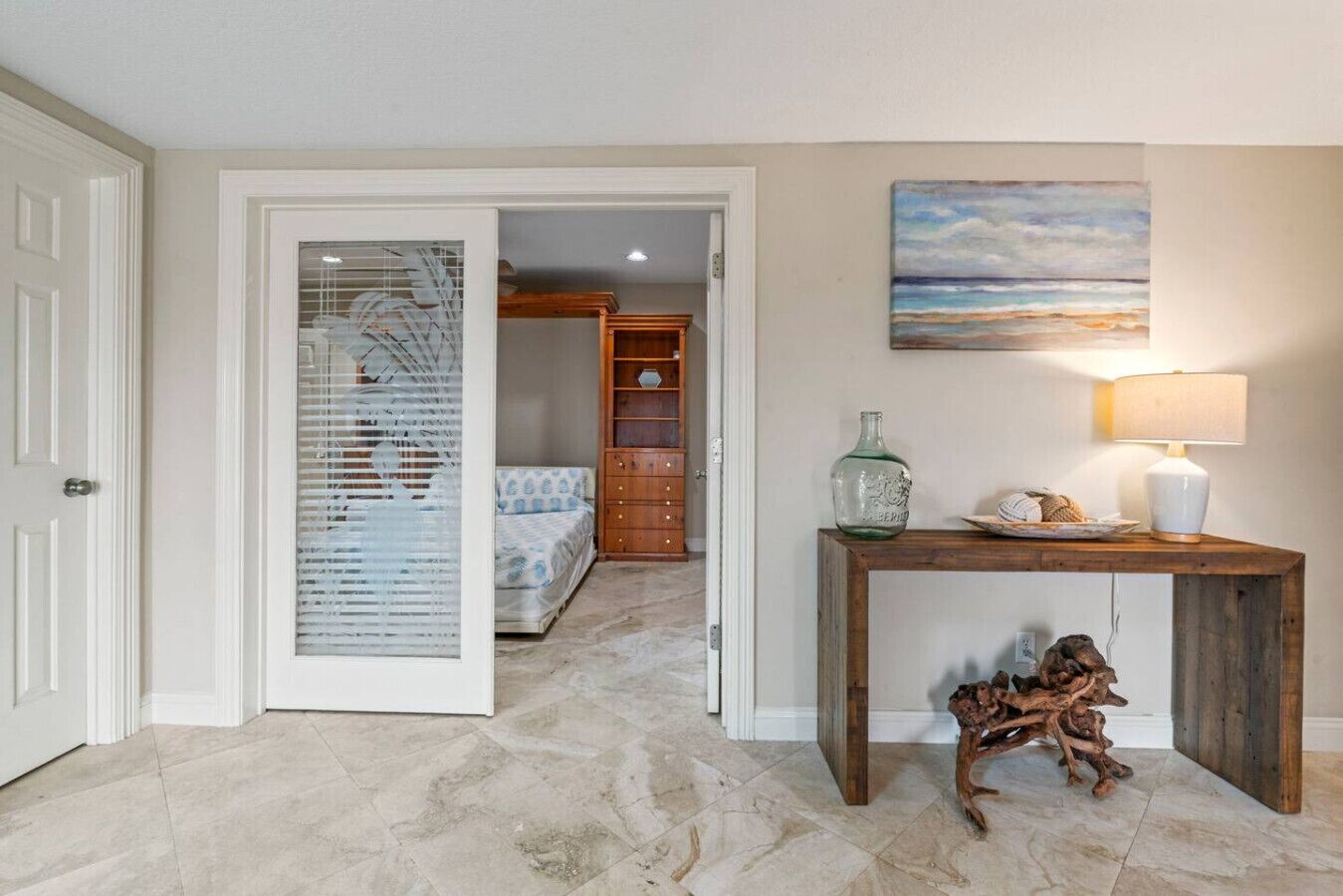 Un pasillo con una cama, una consola de madera con una lámpara, y un cuadro de una playa.