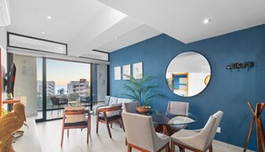 Een moderne woonkamer met een glazen eettafel, comfortabele zitplaatsen, een grote spiegel en een 'WELCOME'-bord aan de muur.