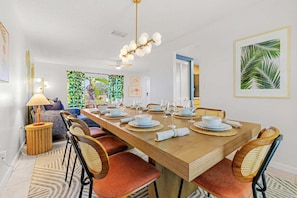 A dining area with a long wooden table set for six, surrounded by wicker chairs, a ceiling fan, and a hanging light fixture.