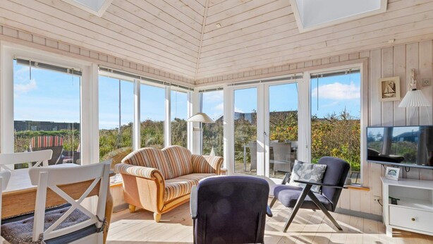 A sunlit room with a high ceiling, large windows, a sofa, chairs, and a TV. - 5 Star Holiday Home in Lokken (Lokken)