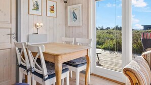 A dining area with a wooden table and white chairs, framed artwork on the wall, and a view of the outdoors through a glass door. - 5 Star Holiday Home in Lokken (Lokken)