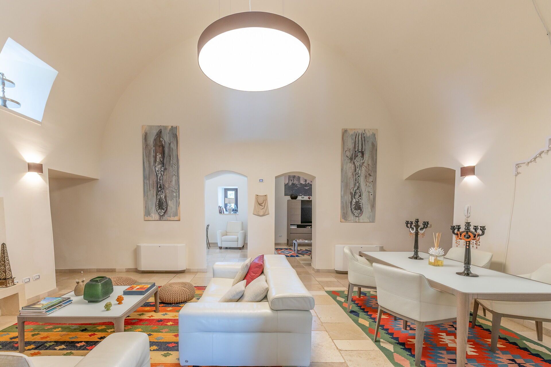 Un ampio soggiorno con un divano bianco, un tavolino da caffè e un'area pranzo con tavolo e sedie. Alle pareti si trovano dipinti astratti e un grande lampadario al soffitto.