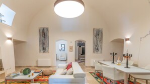 Ein geräumiges Wohnzimmer mit einem weißen Sofa, einem Couchtisch und einem Essbereich mit Tisch und Stühlen. An den Wänden befinden sich abstrakte Gemälde und eine große Leuchte an der Decke.