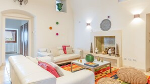Ein geräumiges Wohnzimmer mit einem weißen Sofa, einem Couchtisch und einem farbenfrohen Teppich. An der Wand hängt eine kleine, grüne Vase und über dem Kamin ein dekorativer Spiegel.