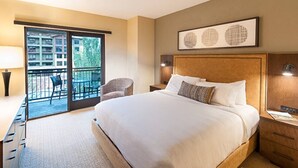 A hotel room with a large bed, a desk, a chair, and a balcony with a view of buildings and trees.