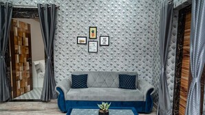 Una sala de estar moderna con un sofá gris, almohadones azules decorativos, una mesa de centro de vidrio con una planta y una pared texturizada con cuadros enmarcados.