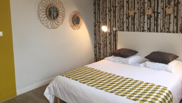 Un lit bien fait, recouvert d’une couvre-lit à motifs, deux miroirs décoratifs fixés au mur, et un mur orné d’un papier peint à motifs verticaux et de décorations en forme de nichoir.