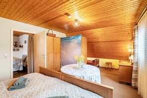 A bedroom with a wooden ceiling, a bed with a rolled towel, a small round table with a vase of flowers, and a wooden cabinet.