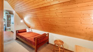 Une petite pièce agréable, dotée d’un plafond en bois incliné, d’un lit simple avec un matelas rouge, d’un petit tabouret et d’une salle de bain visible, équipée d’une douche.