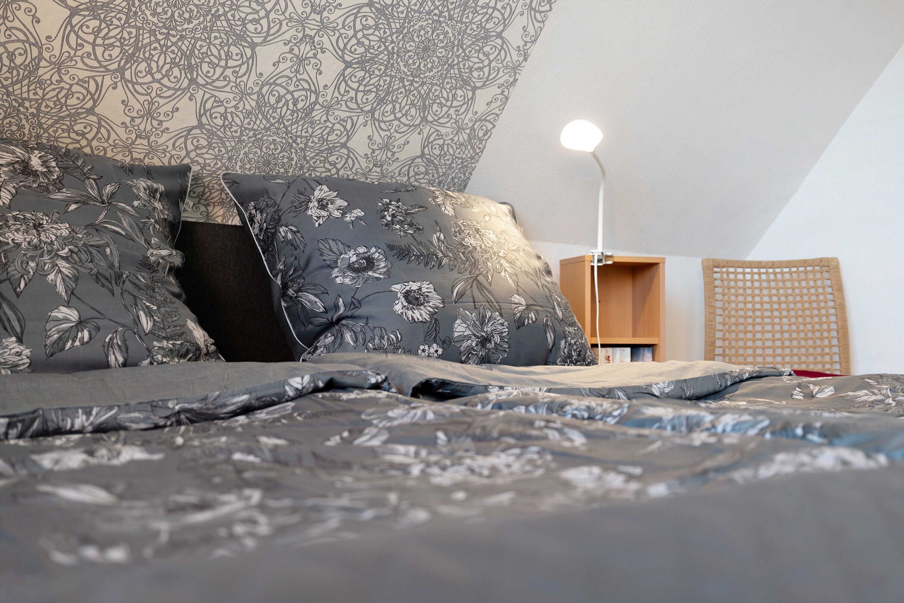 A neatly made bed with floral-patterned pillows and a grey bedspread, a bedside lamp, a wooden nightstand, and a woven basket.