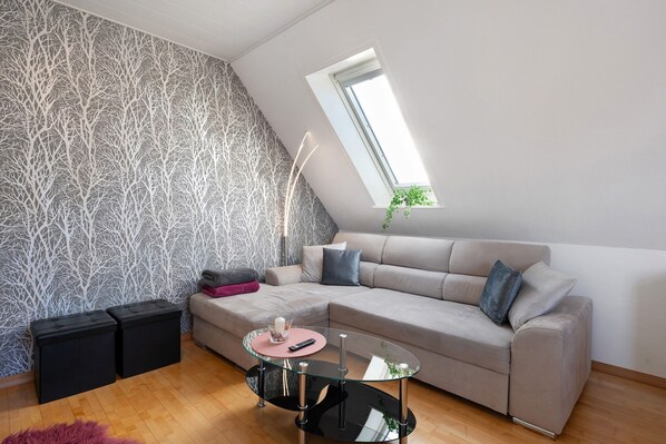 A modern living room with a sectional sofa, a glass coffee table, black ottomans, a patterned wallpaper, a skylight, and a small plant.