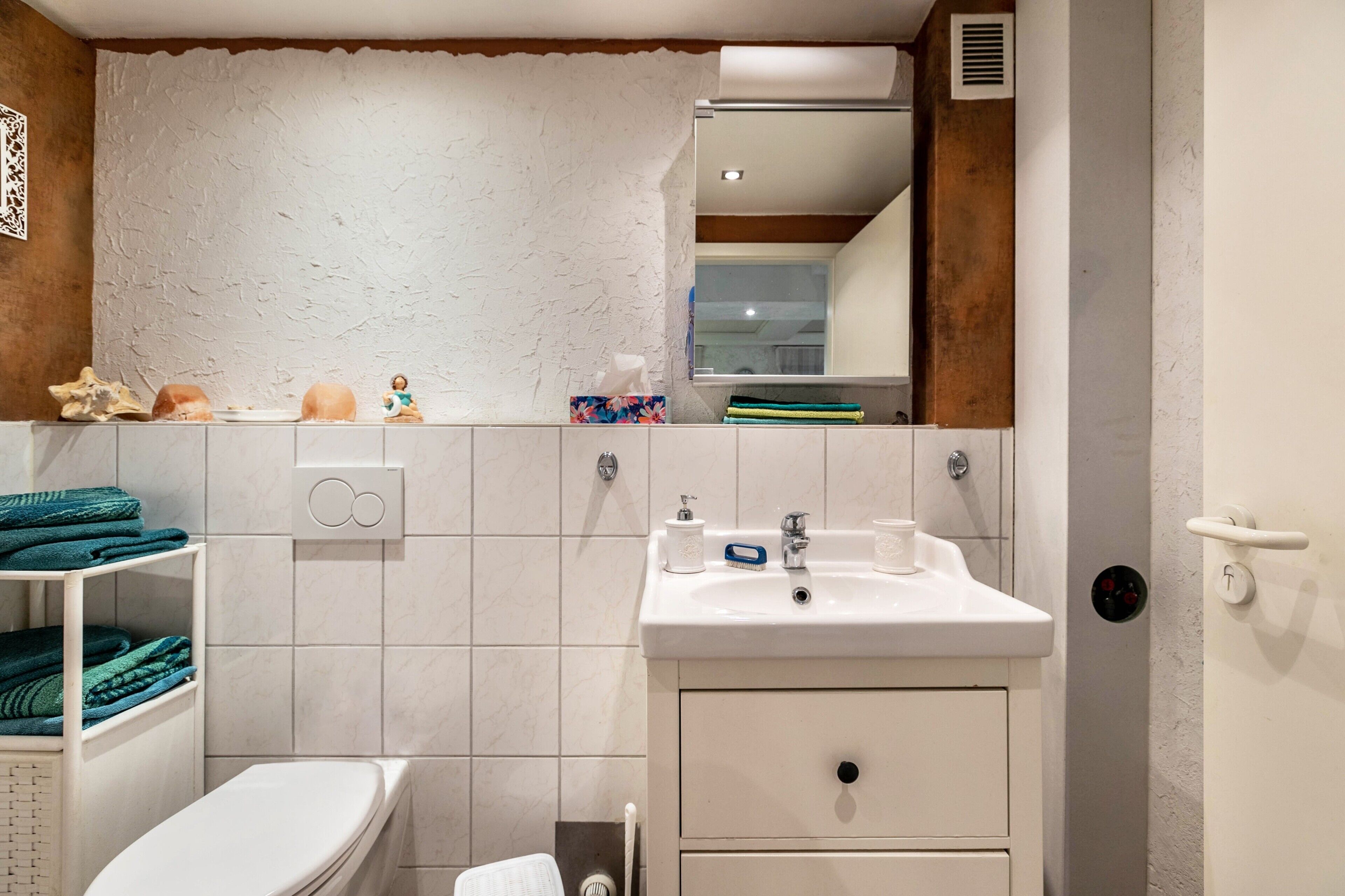 A bathroom with a white sink, mirror, and toilet. There are towels on a shelf and a decorative item on the wall.