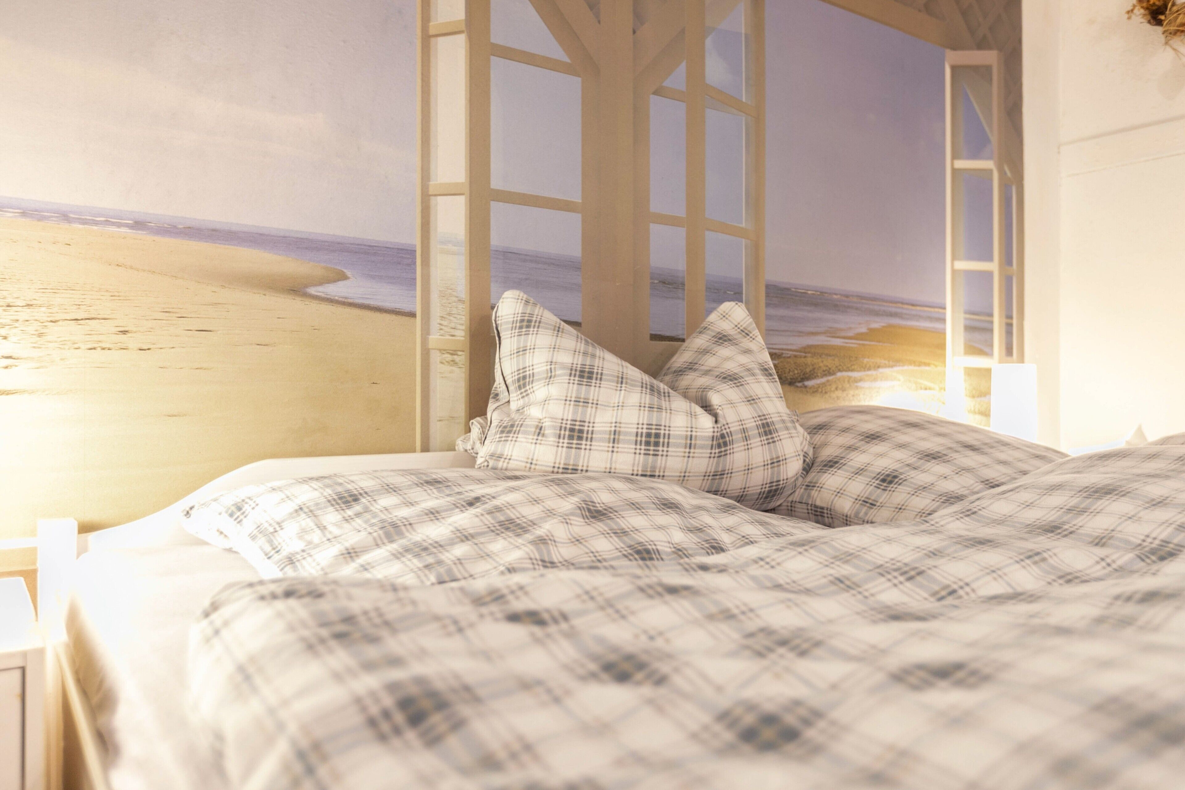 A bed with a checkered bedspread, a window with a view of a beach, and a ladder leading to a balcony.