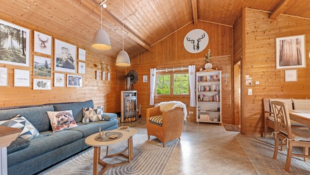 Un intérieur en bois comprenant un canapé, un fauteuil en osier, une table, une bibliothèque et des tableaux encadrés accrochés aux murs.
