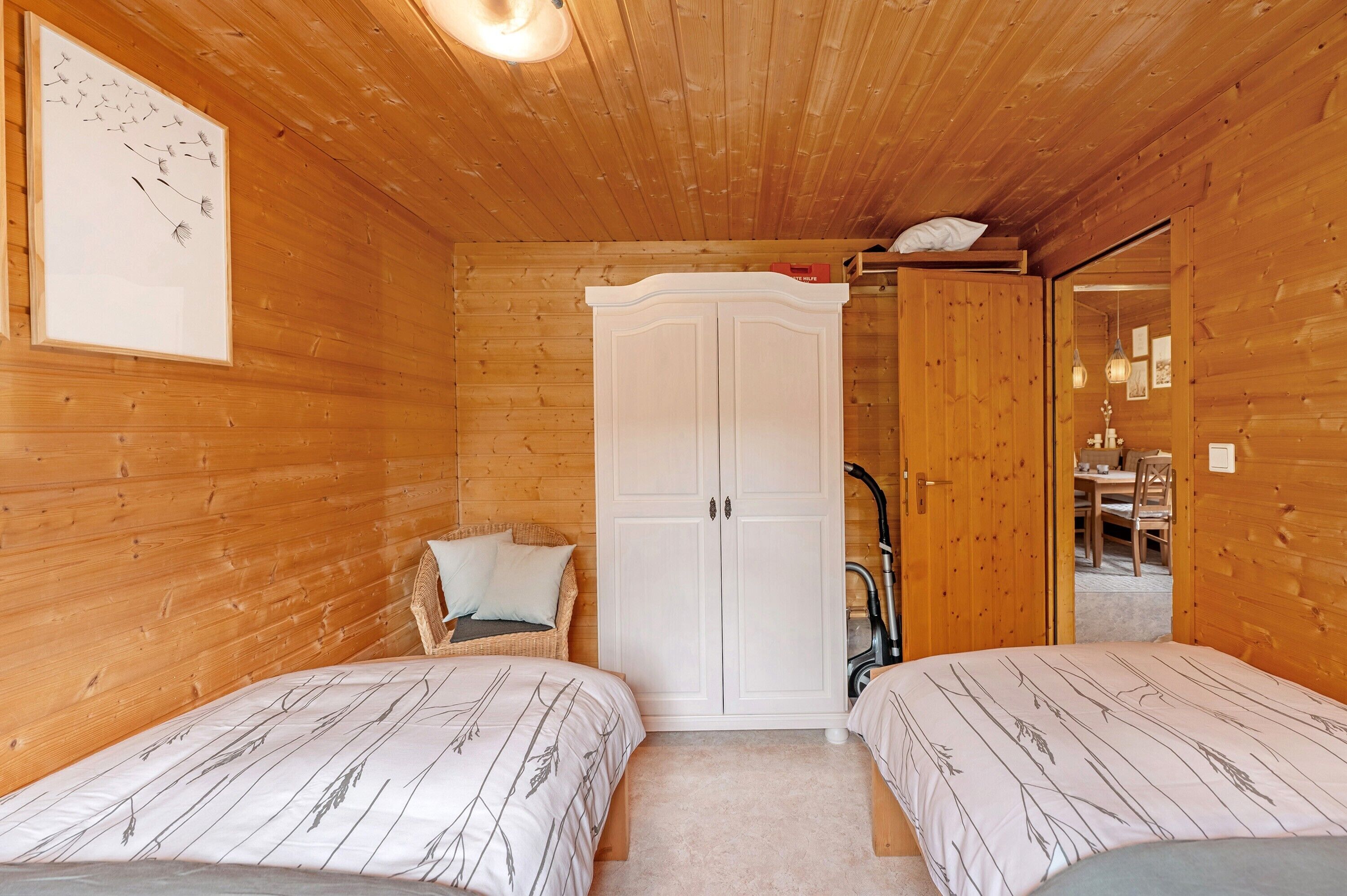 A wooden cabin room with two beds, a white wardrobe, and a dining area visible through an open door.