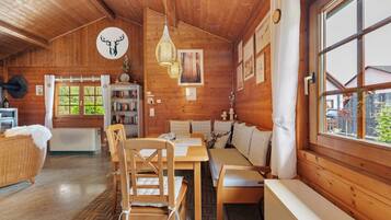 A cozy cabin interior with wooden walls, a dining area, a sofa, and a window with a view.