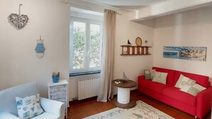 Una sala de estar con un sofá rojo, un sillón blanco, una mesita, un radiador, una ventana con cortinas y decoraciones en la pared, incluyendo un adorno con forma de corazón y una estantería con una raqueta de tenis.