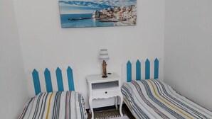 Un dormitorio con dos camas individuales, una mesita de noche, una lámpara y un cuadro de un pueblo costero.