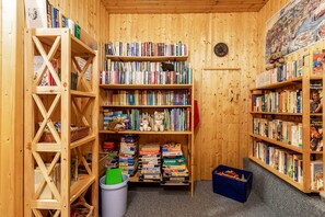 Une pièce aux murs lambrissés, avec des étagères remplies de livres et de jouets, une poubelle et un bac de rangement bleu.