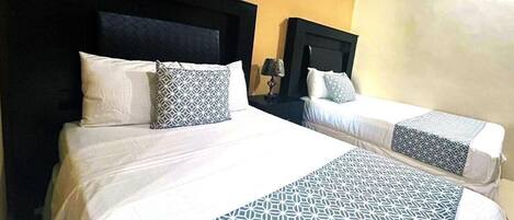 A hotel room with two beds, each with a patterned bedspread and pillows. There is a nightstand with a lamp between the beds.