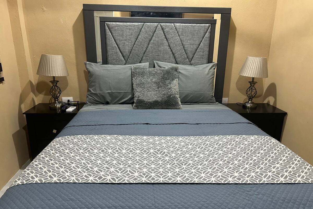Una cama bien hecha con una colcha de diseño geométrico, un edredón acolchado y dos almohadas. Hay dos mesitas de noche con lámparas y un cabecero fijado a la pared.
