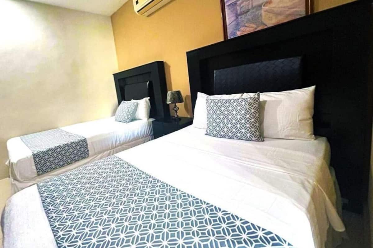 Habitación de hotel con dos camas, una cama grande con cubrecama de estampado geométrico y dos camas más pequeñas con cubrecamas estampadas.