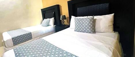 A hotel room with two beds, a large bed with a geometric patterned bedspread and two smaller beds with patterned bedspreads.