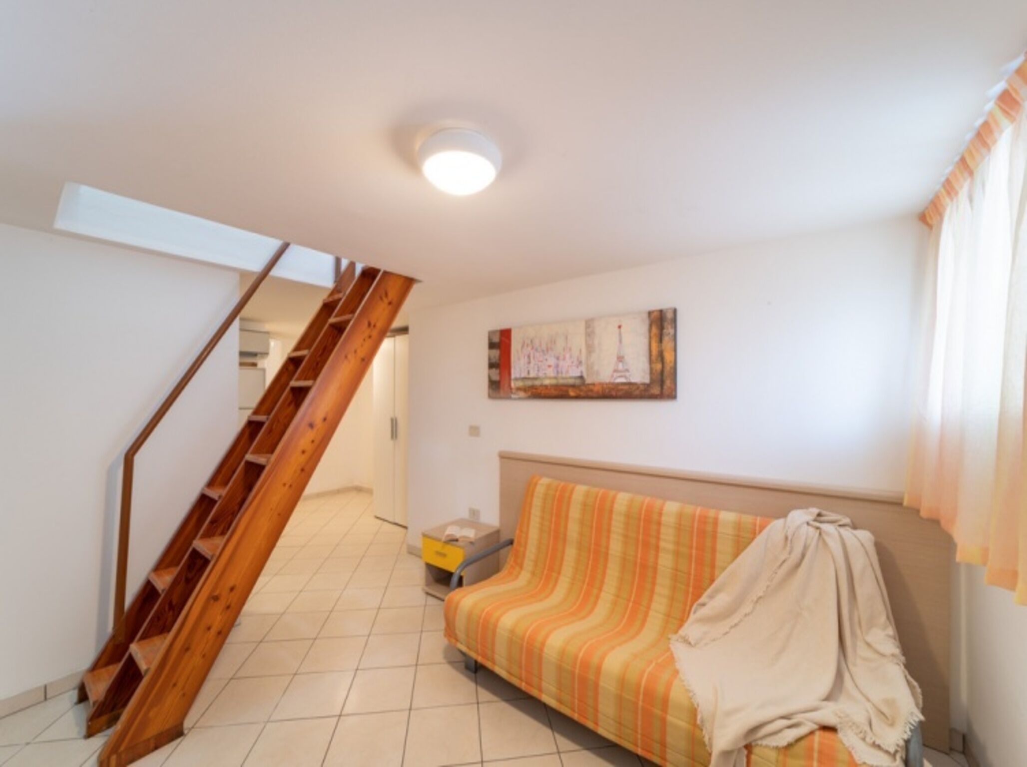 Quarto com escada de madeira, cama com manta listrada e pintura de uma paisagem urbana.