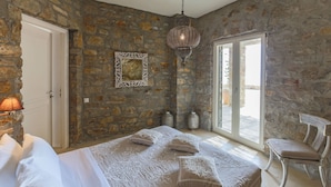 A bedroom with a stone wall, a bed with ornate bedding, a hanging lamp, and a view through a glass door. - Villa in Kalafati with 8 bedrooms sleeps 16 (Kalafati)