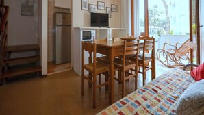 Ein kleines Zimmer mit einem Bett, einem Esstisch mit Stühlen, einer Küchenzeile inklusive Kühlschrank und Mikrowelle, einem Fernseher und einem Fenster mit Blick nach außen.