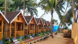 A row of wooden cabins with pitched roofs, surrounded by palm trees. - Sand Tent Beach Resort Patnem (Canacona)