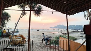 A beach view from a covered area with a thatched roof, palm trees, and a sandy beach with people and boats. - Sand Tent Beach Resort Patnem (Canacona)