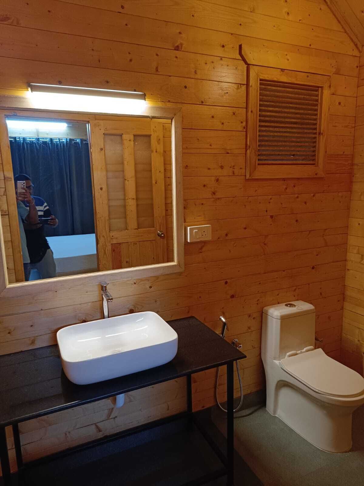 Baño de cabaña de madera con lavamanos blanco, espejo, inodoro y ventana con contraventana.