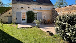Une maison en pierre avec un toit en pente, une cheminée et un petit jardin agrémenté d’une table et de chaises.