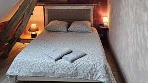 Une chambre avec un plafond en pente, un lit recouvert d’une couvre-lit à motifs floraux, deux lampes de chevet et une structure en poutres apparentes.