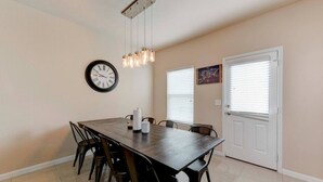 Un coin repas comprenant une table rectangulaire, six chaises, une horloge murale, des luminaires suspendus et une porte avec des stores.