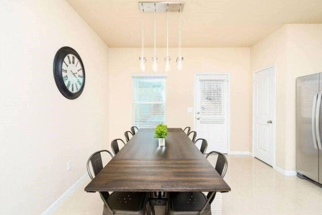 A dining room with a long table, chairs, a clock, and a refrigerator.