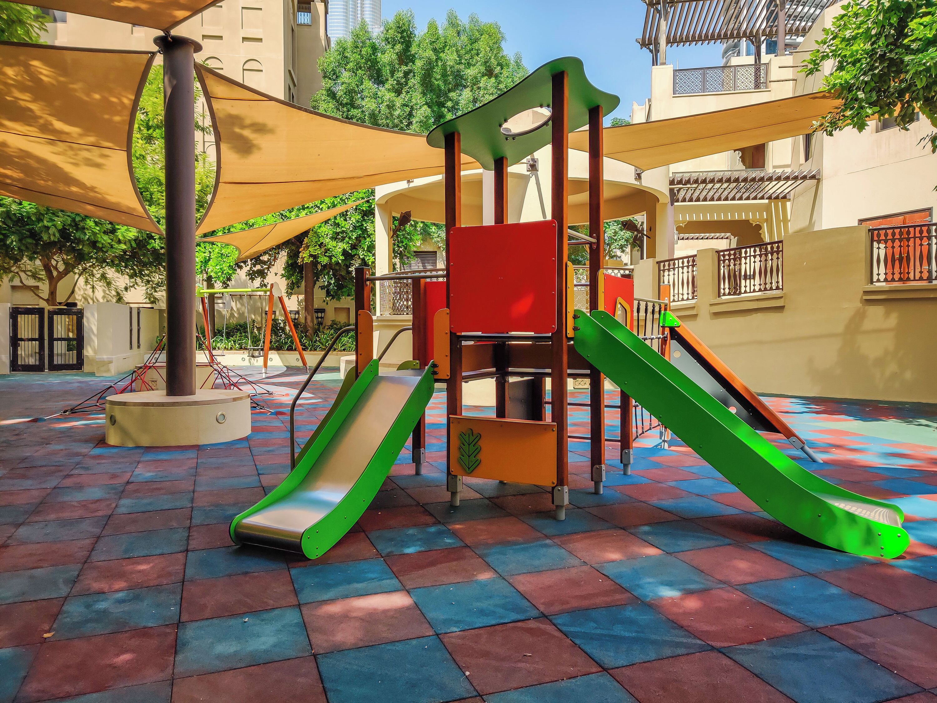 Un espace de jeux pour enfants avec des toboggans et des structures d’escalade, entouré d’une zone carrelée et ombragé par une marquise.