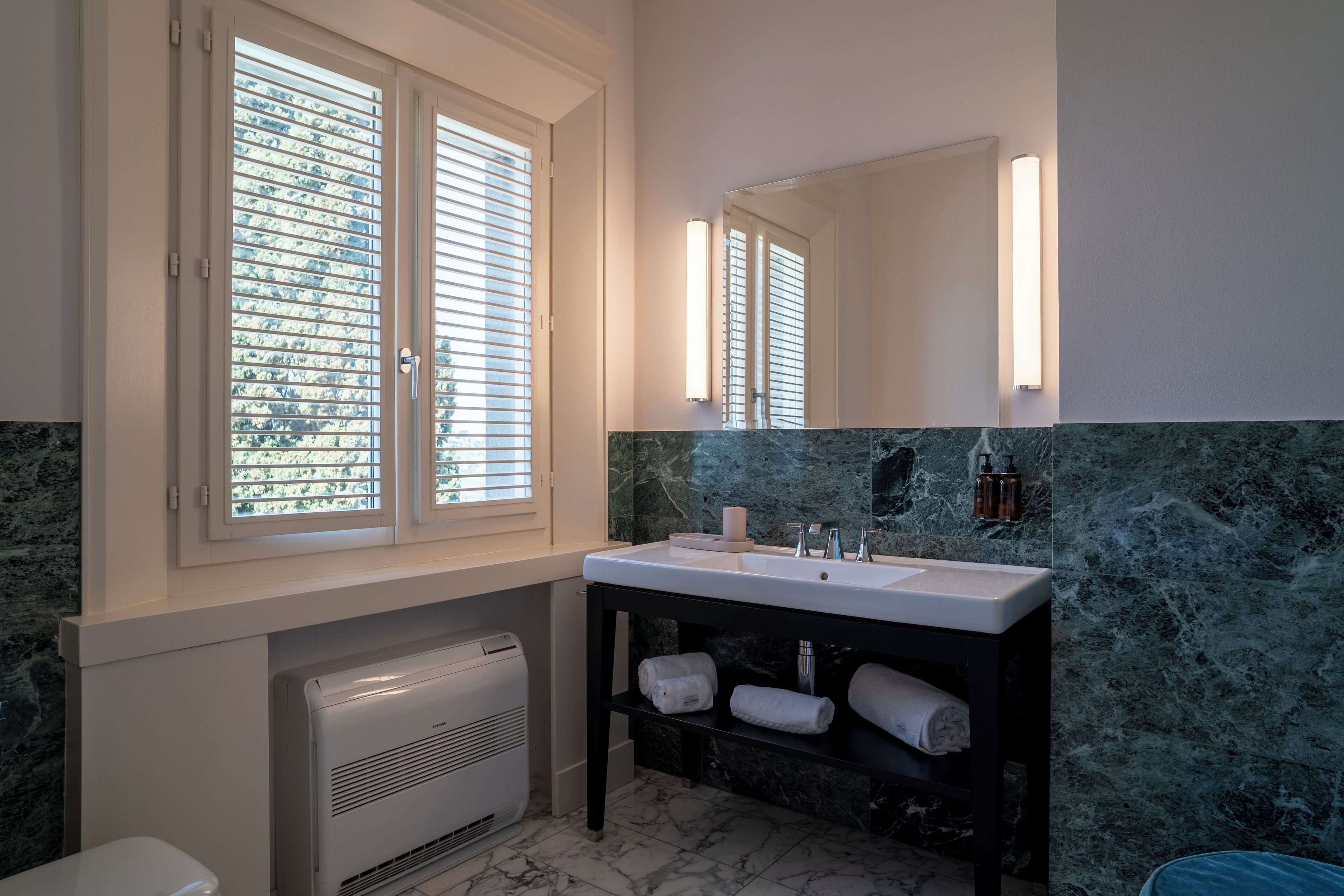 Une salle de bain avec un lavabo blanc, un grand miroir et une fenêtre avec des stores.
