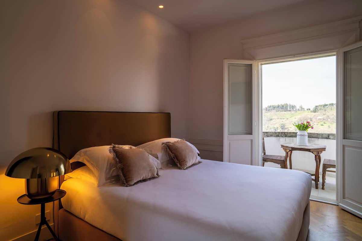 A bedroom with a large bed, a bedside lamp, and a view of a balcony with a table and chairs.