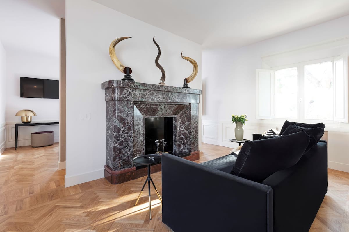 A modern living room with a marble fireplace, a black sofa, and a wooden herringbone floor.