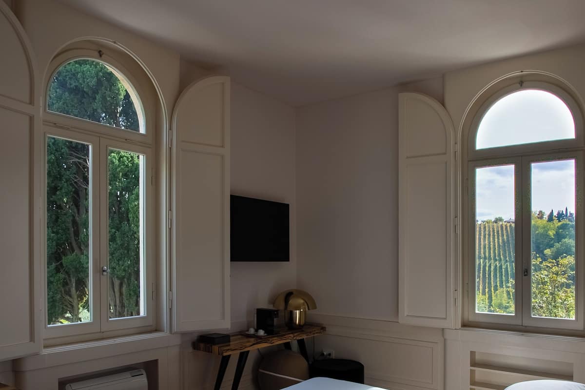 A bedroom with a large bed, two windows with arched tops, a flat-screen TV on the wall, and a desk with a lamp.