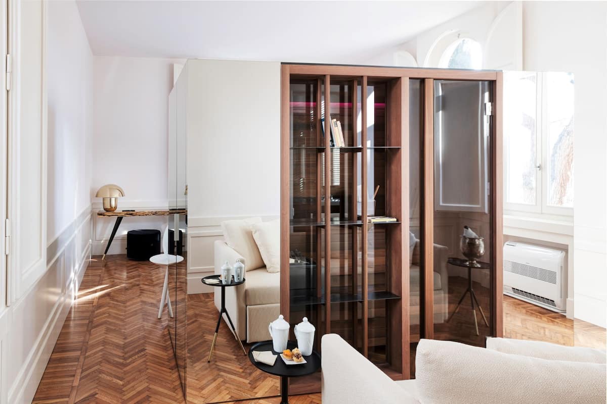 A modern living room with a herringbone wood floor, a white sofa, a glass-fronted bookcase, and a small round table with a lamp and a tray of pastries.