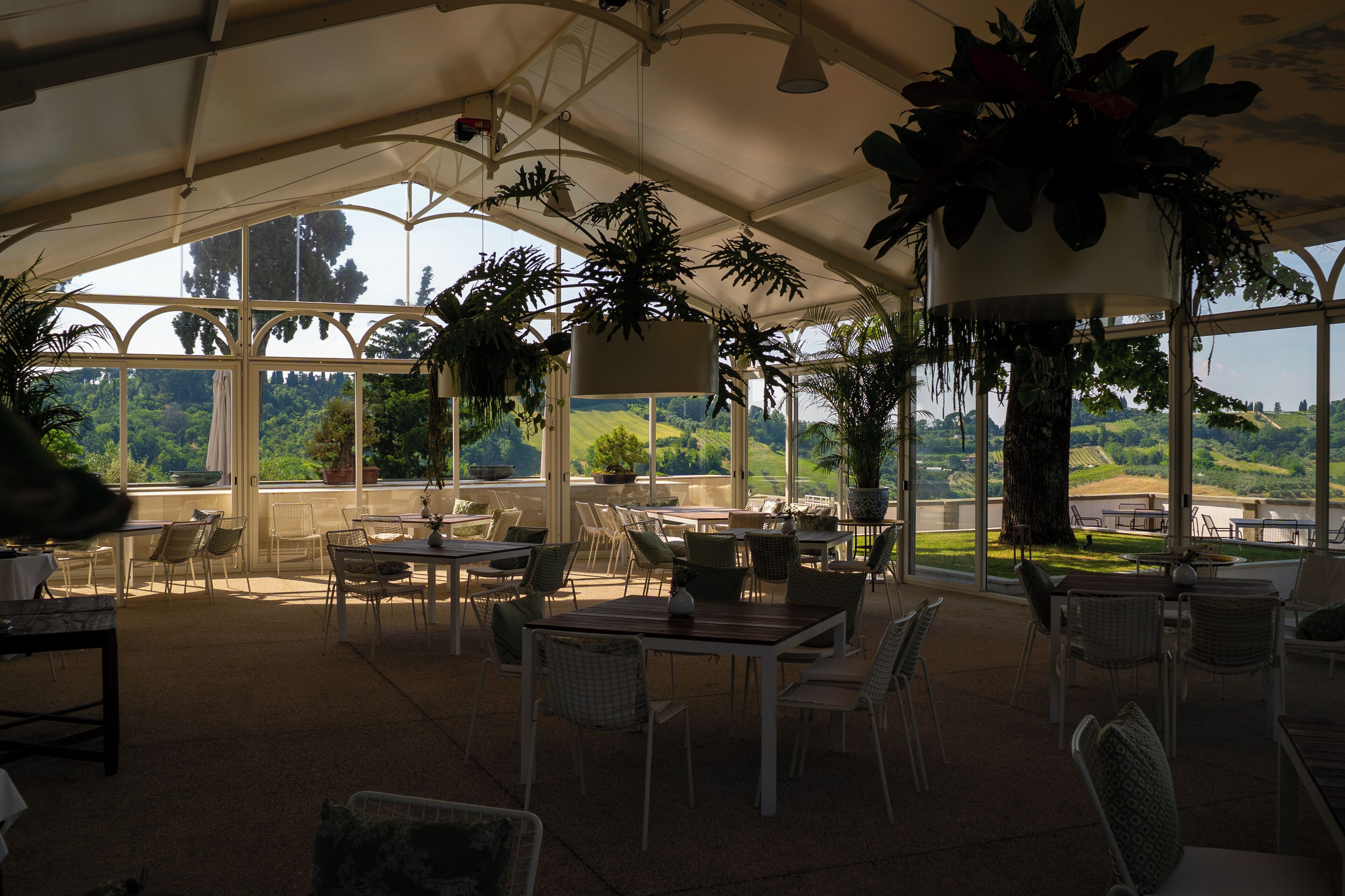 A covered outdoor seating area with tables and chairs, surrounded by large windows offering a view of the countryside.