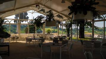 A covered outdoor seating area with tables and chairs, surrounded by large windows offering a view of the countryside.