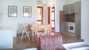 A dining area with a table and chairs, a patterned armchair, a kitchen with cabinets and appliances, and two framed pictures on the wall. - Apartment in Porto Pollo Near Sea Beach (Palau)