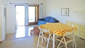 A room with a dining table, chairs, a sofa, a small cabinet, and a sliding glass door leading to an outdoor area. - Apartment in Porto Pollo Near Sea Beach (Palau)