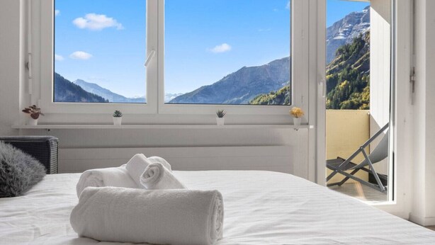 A bedroom with a bed, a window with a mountain view, a chair outside, and a small plant on the windowsill. - Charming studio near the ski slopes (Leukerbad)