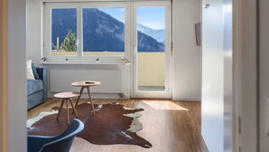A room with a wooden floor, a cowhide rug, a blue sofa, a small round table, and a view of mountains through large windows. - Charming studio near the ski slopes (Leukerbad)